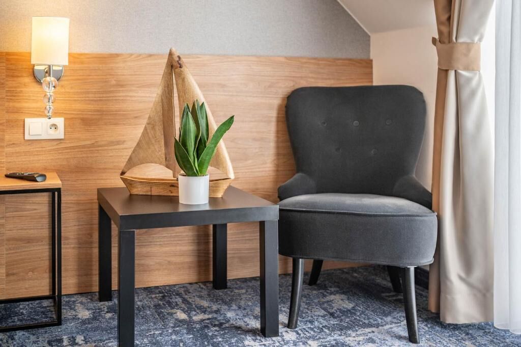 Et hotelværelse med en mørkegrå lænestol, et lille træsidebord med en potteplante, en lampe og en fjernbetjening.