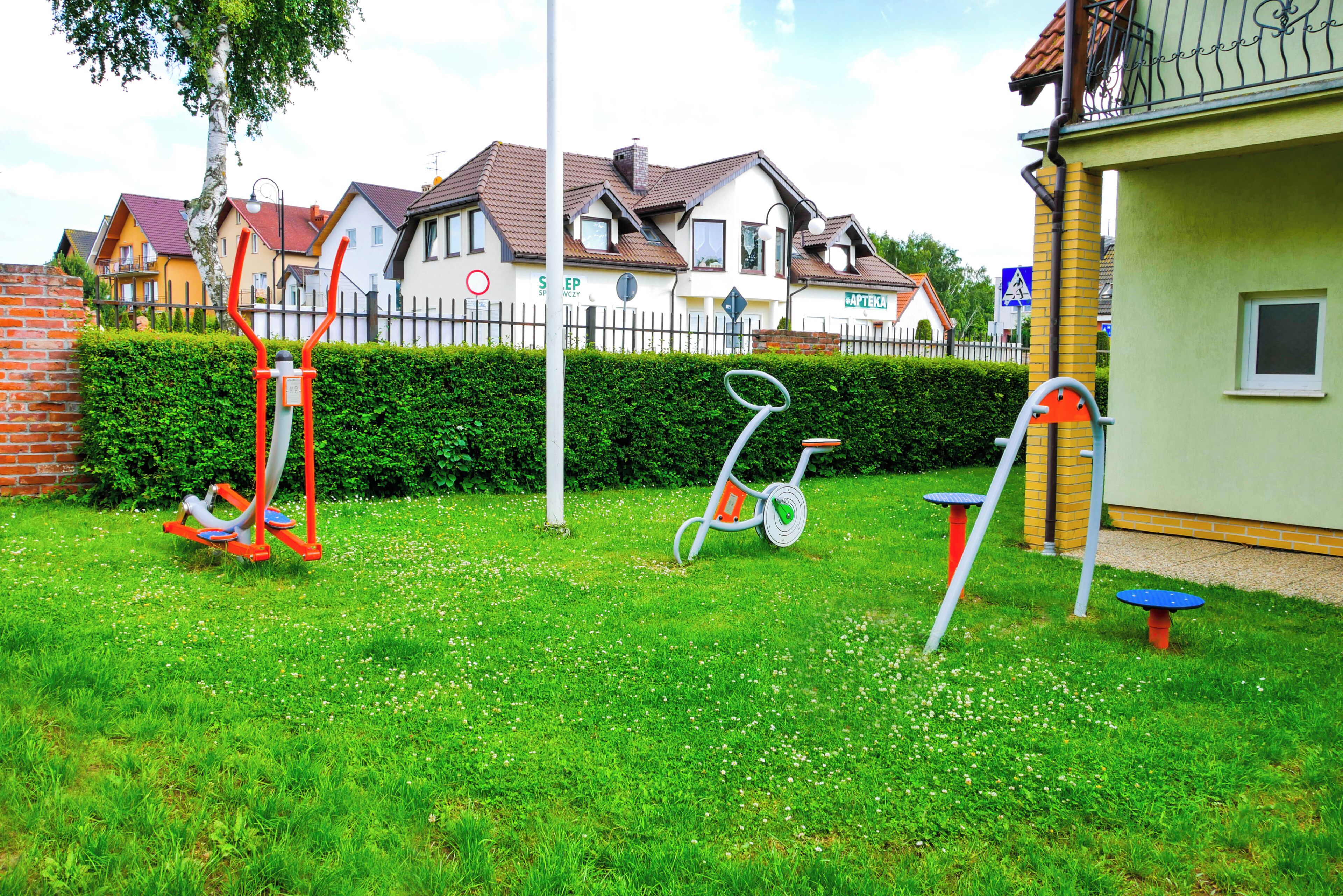 A grassy area with outdoor exercise equipment, a hedge, and a row of houses in the background.