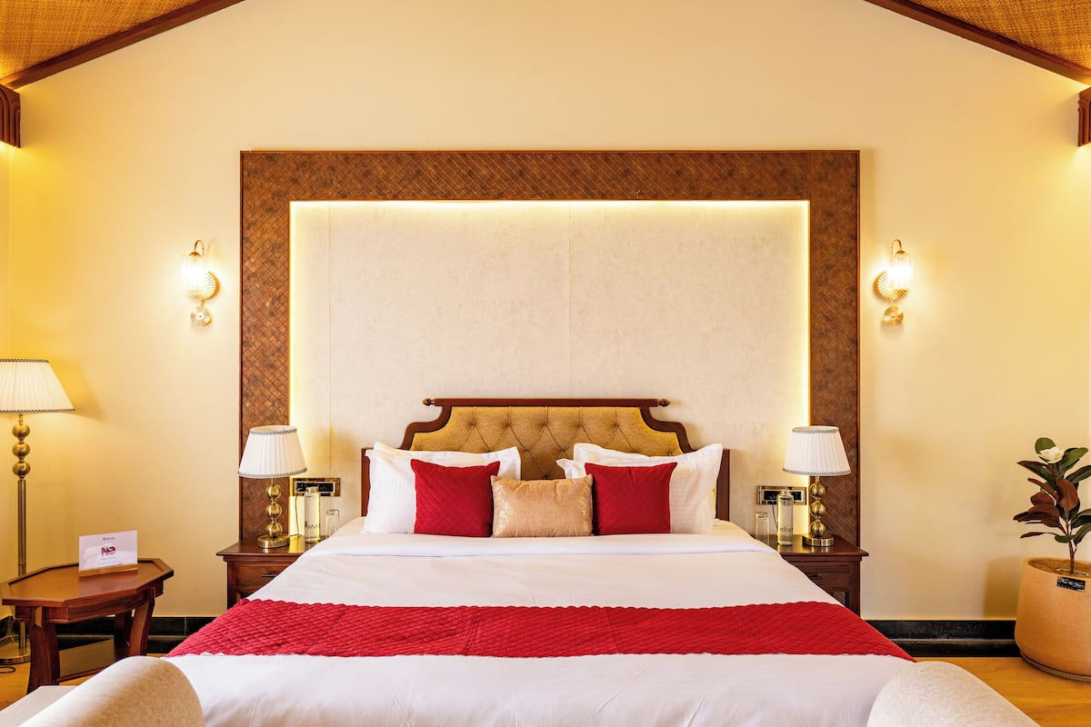 A hotel room with a large bed, two bedside tables, a nightstand, a red throw blanket, and wall sconces.