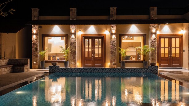 Une piscine avec un bord carrelé, un mur en pierre et un bâtiment doté de nombreuses portes et de plusieurs luminaires.