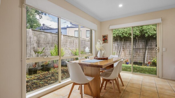 Un comedor con mesa de madera, sillas blancas y ventanas amplias con vista a un patio trasero con valla de madera y plantas.