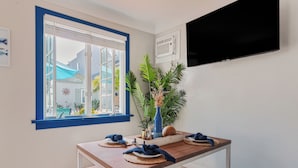 Ein Essbereich mit Holztisch, Fensterrahmen in Blau, Topfpflanzen und einem Wand-Fernseher.