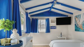 Ein Zimmer mit Bett, Fernseher, kleinem Tisch mit Flasche und Gläsern, einem Fenster mit Vorhängen und einer Pflanze.