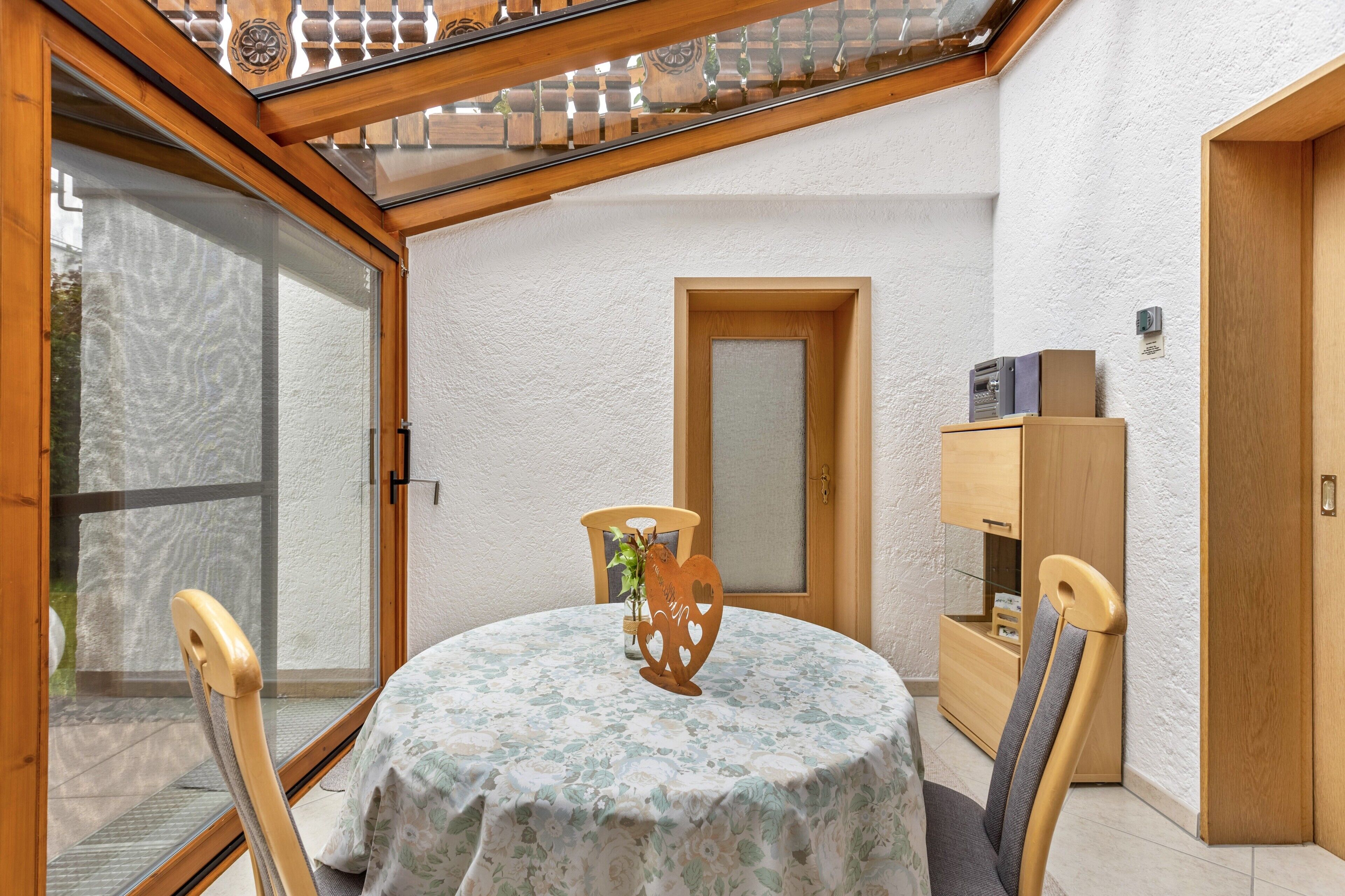 Ein runder Tisch mit einer blumigen Tischdecke, eine Vase mit herzförmigem Design und zwei Holzstühle.