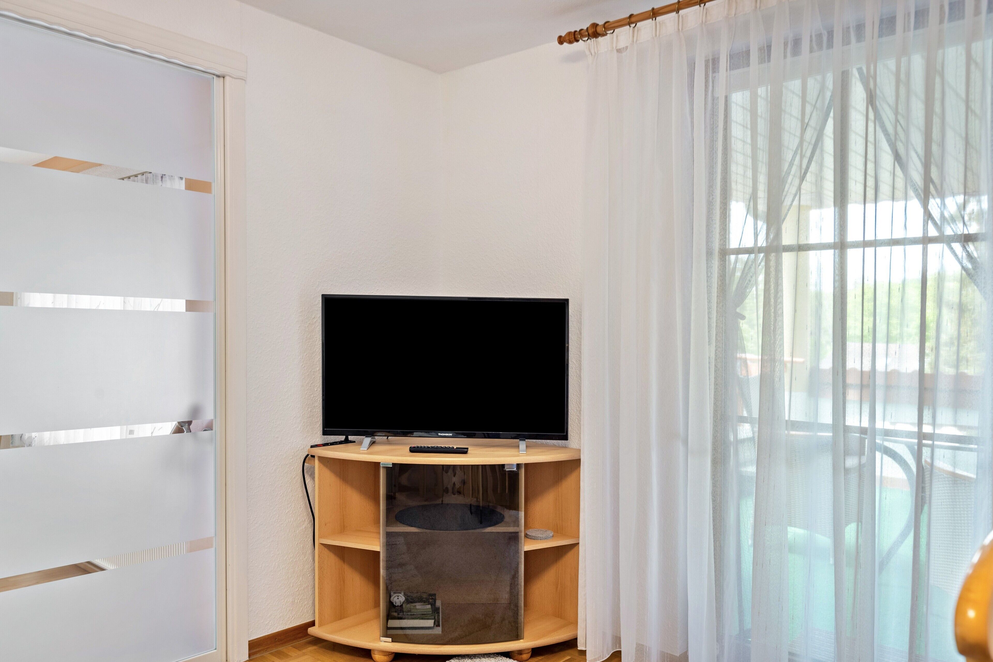 Une pièce avec un téléviseur posé sur un meuble en bois, une étagère en verre, une porte avec des panneaux en verre dépoli et une fenêtre ornée de rideaux légers.