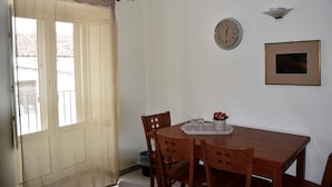 Een eetgedeelte met een houten tafel, vier stoelen, een klok, een lijst met een afbeelding en een raam met gordijn.