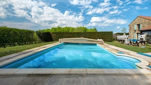 Une piscine rectangulaire entourée d’une terrasse en pierre, elle-même bordée d’une haie et d’une maison dotée d’un toit en tuiles et d’une porte en verre.