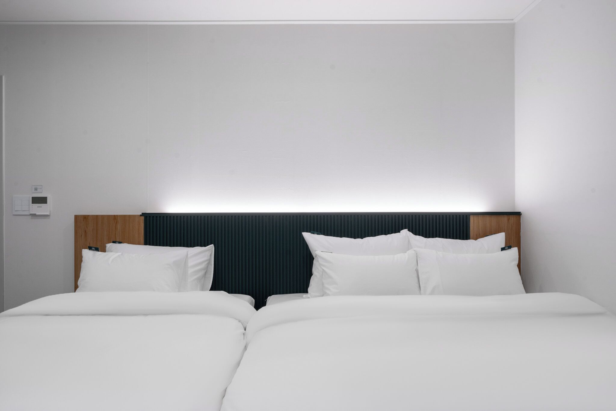 Una cama doble con cabecera de madera, ropa de cama blanca y un termostato fijado a la pared.