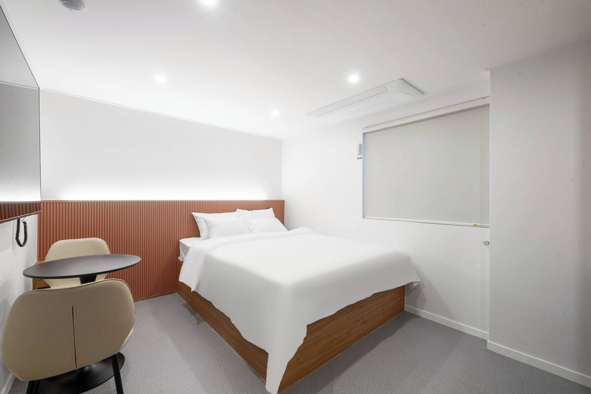 Ein Hotelzimmer mit Bett, einem kleinen Tisch, einem Stuhl, einer Whiteboard-Tafel und einem Holz-Kopfteil.
