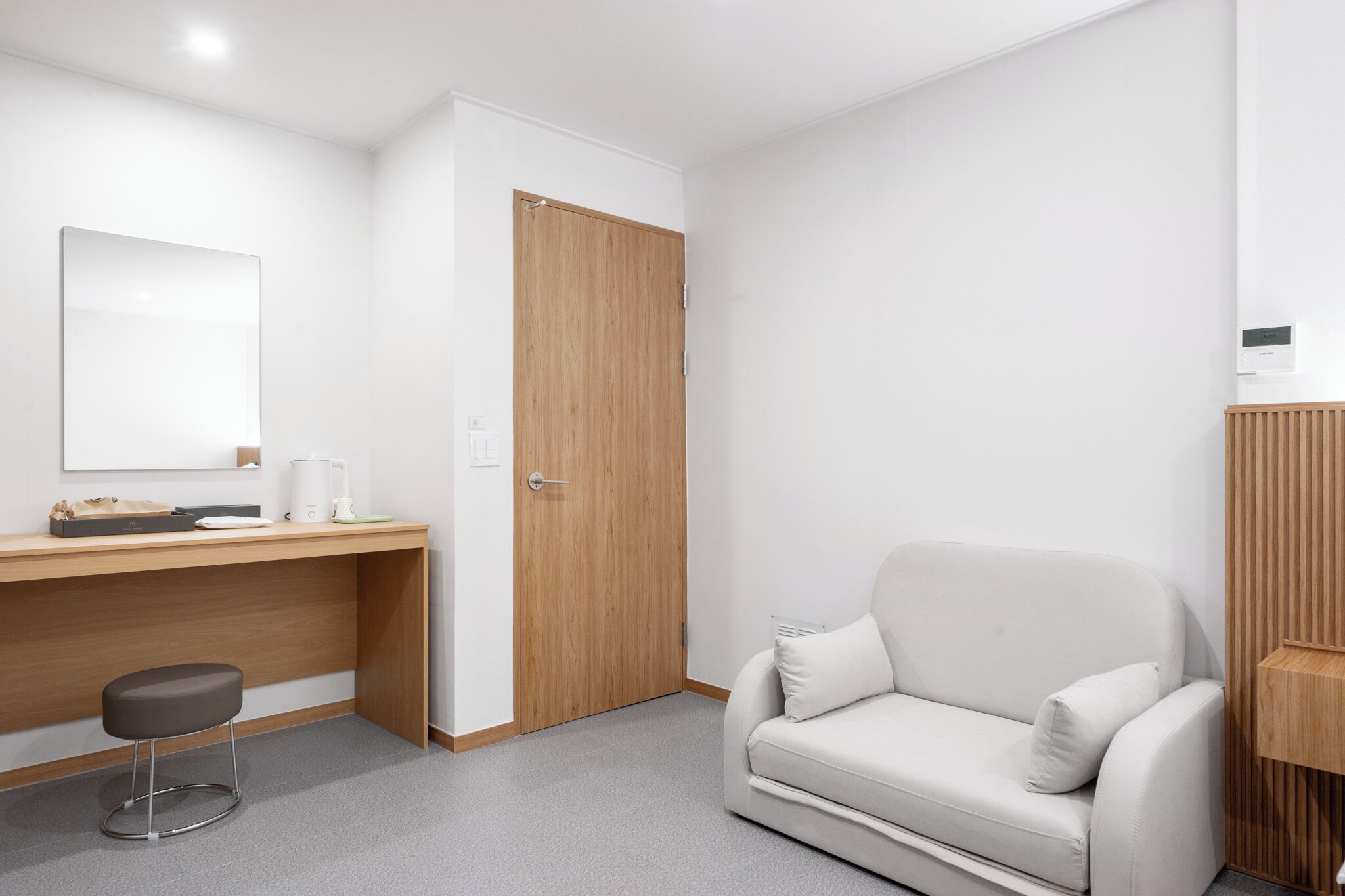 A modern hotel room with a wooden desk, a white sofa, a small stool, and a closed wooden door.