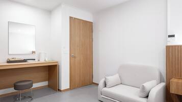 A modern hotel room with a wooden desk, a white sofa, a small stool, and a closed wooden door.