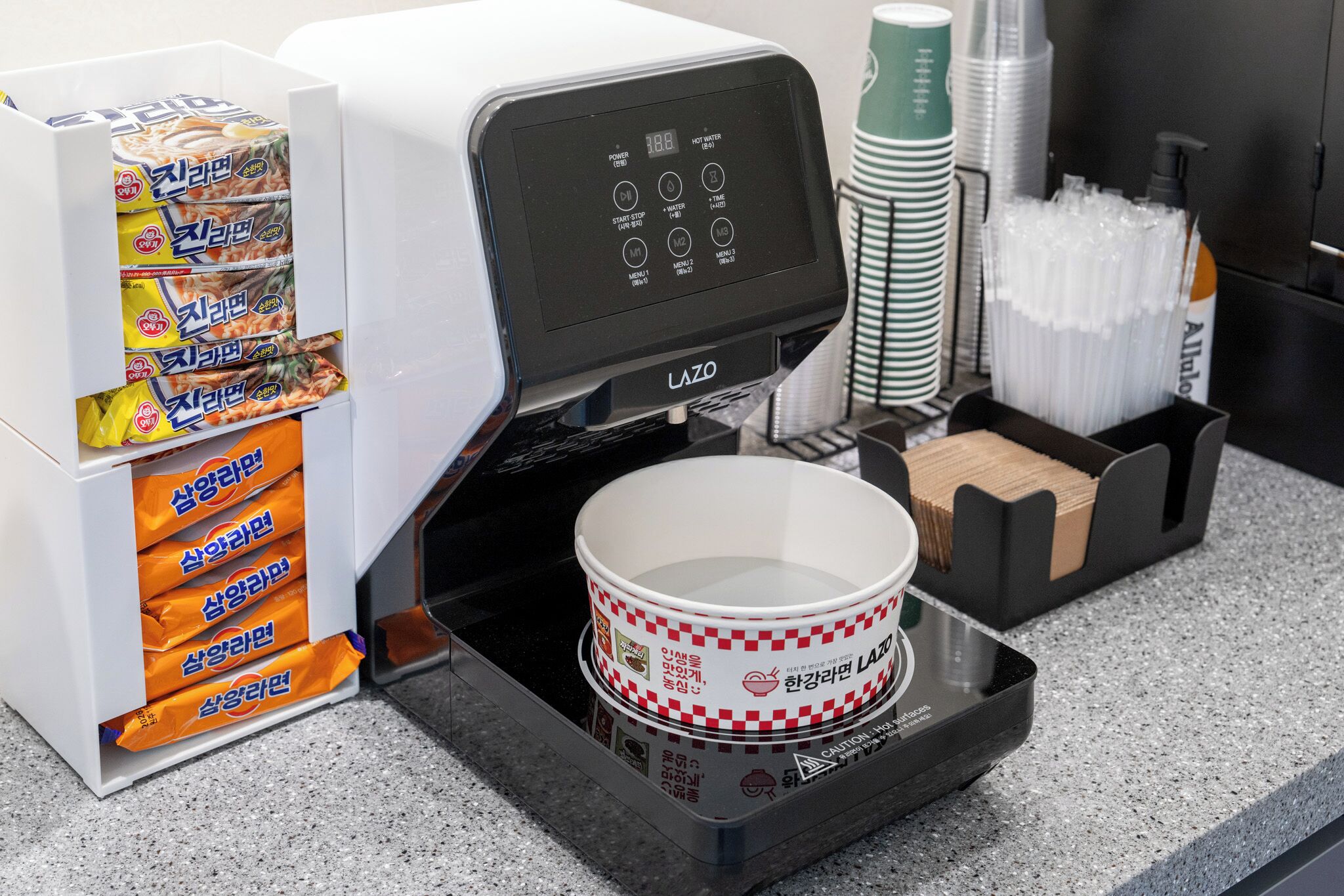 Une machine à café dotée d’un écran numérique, une pile de gobelets jetables et un plateau avec un bol.