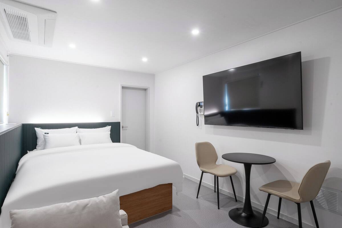 Ein Hotelzimmer mit Bett, einem an der Wand montierten Fernseher, einem kleinen Tisch mit zwei Stühlen und einer Tür.