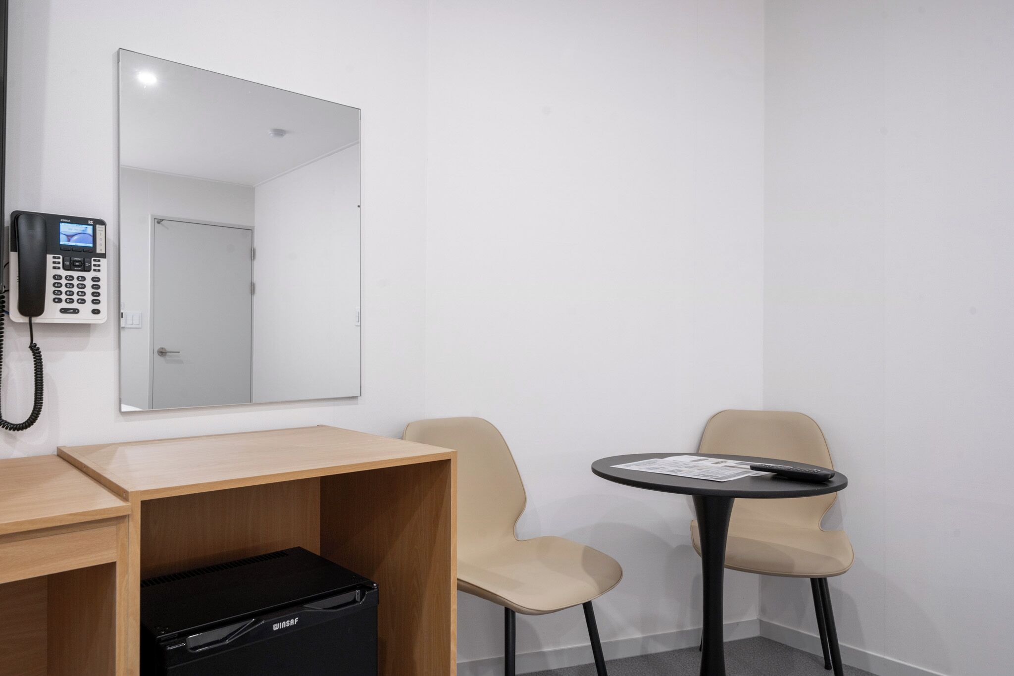 A room with a desk, two chairs, a small table, a black briefcase, a phone, and a mirror.