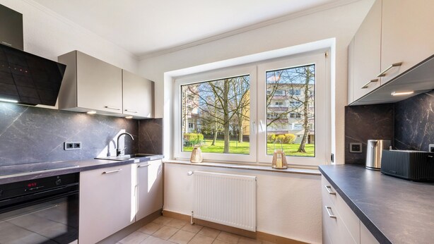 Una cocina moderna con horno empotrado, una ventana con vistas a un parque verde y un radiador blanco.