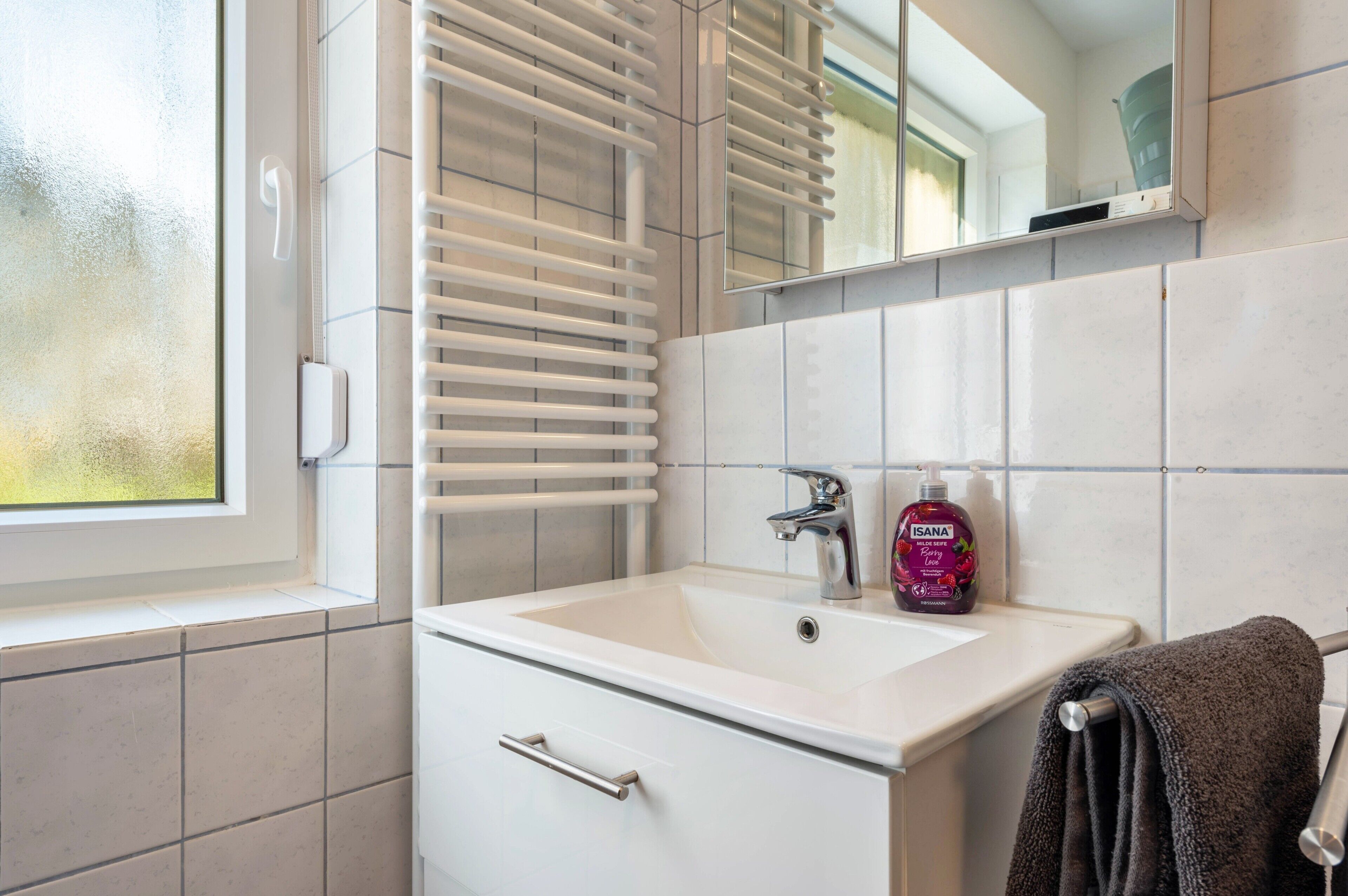 Ein Badezimmer mit einem weißen Waschbecken, einem Spiegel, einem Handtuchhalter und einem Fenster.