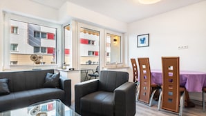 Una sala de estar moderna con sofá, sillón, mesa de centro de vidrio, mesa de comedor con sillas y ventanas grandes con vista a un edificio.