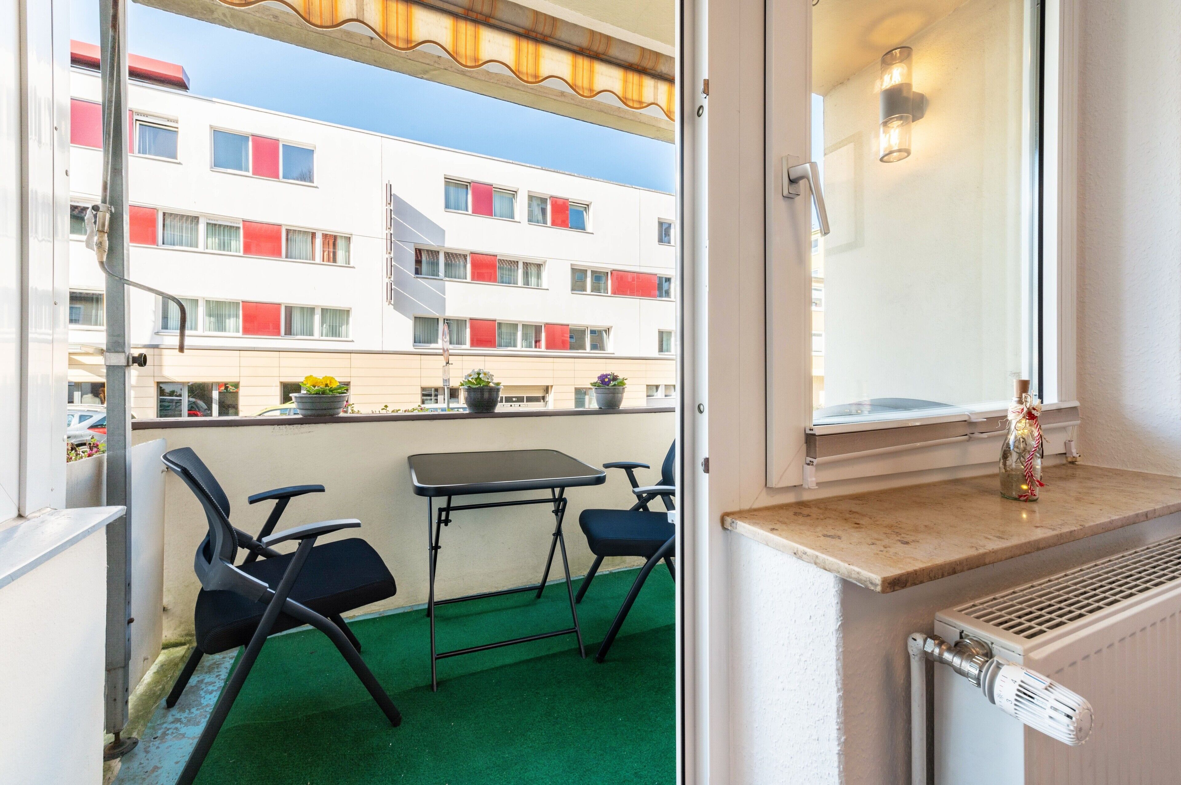 A balcony with a table and chairs, a radiator, and a view of a building with red window shutters.