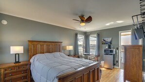 A bedroom with a wooden bed, two bedside tables with lamps, a ceiling fan, a view through a window, and a kitchenette area. - Bagnell Strip Studio for Shootout Weekend Fun (Lake Ozark)