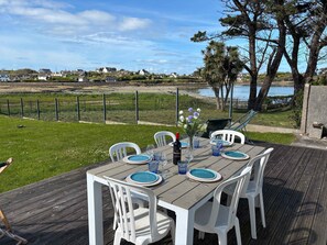 Un área de comedor al aire libre con una mesa preparada para cuatro, rodeada de sillas blancas, con vistas a una masa de agua y una zona vallada con casas al fondo.