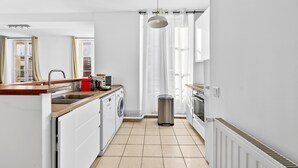 Una cocina moderna con lavadora incorporada, un cubo de basura y una ventana con cortinas de gasa.