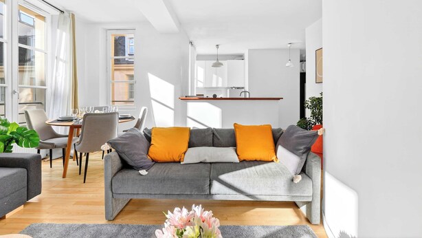 Una sala de estar moderna con un sofá gris, cojines naranjas, una mesa de comedor y una mesa de centro con flores.
