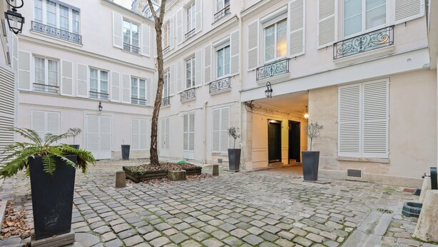 A courtyard with cobblestone paving, white shutters, and a black planter with a fern. - Quartier Palais Royal - Studio Apartment NetFlix (Paris)