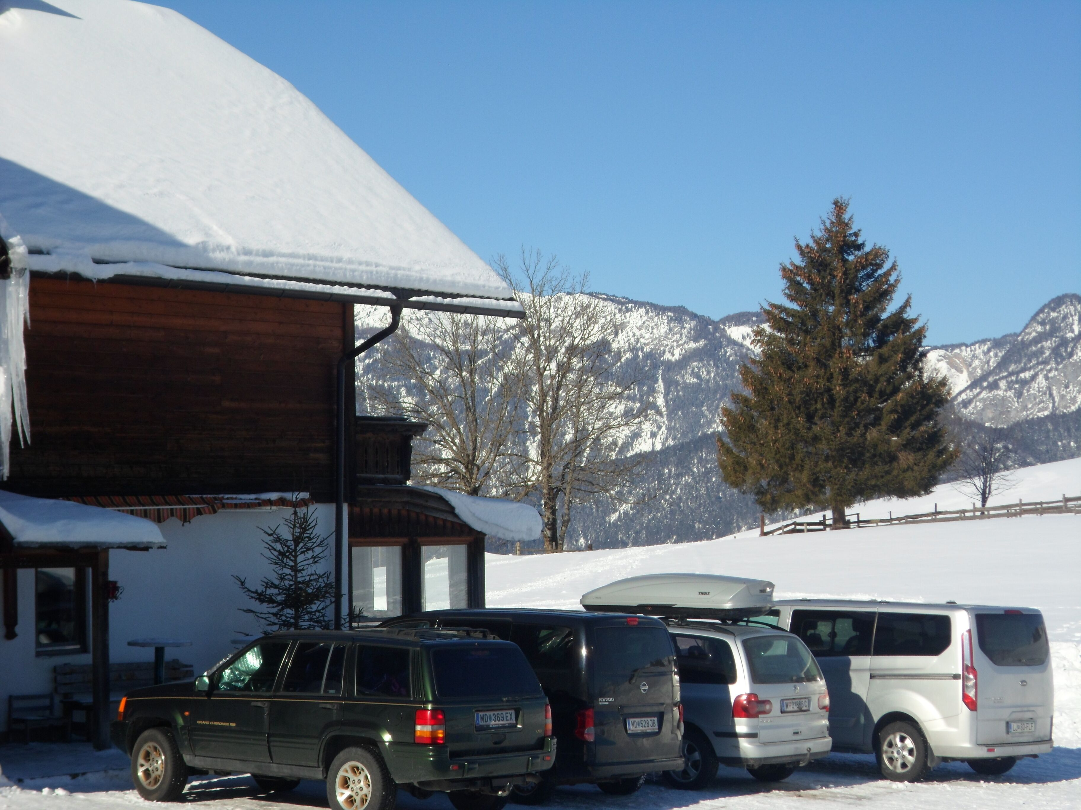 A building with a sloped roof, parked vehicles, and snow-covered mountains in the background.