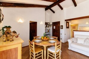 A dining area with a wooden table set for four, surrounded by chairs. There is a white sofa to the right and a kitchen area with a wooden countertop. - Casa Urania Apartment with Shared Pool, Private Terrace, and Wi-Fi (Liscia di Vacca)