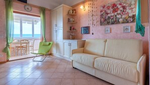 A living room with a sofa, a dining table with chairs, a green chair, and a kitchen area with cabinets and a painting.