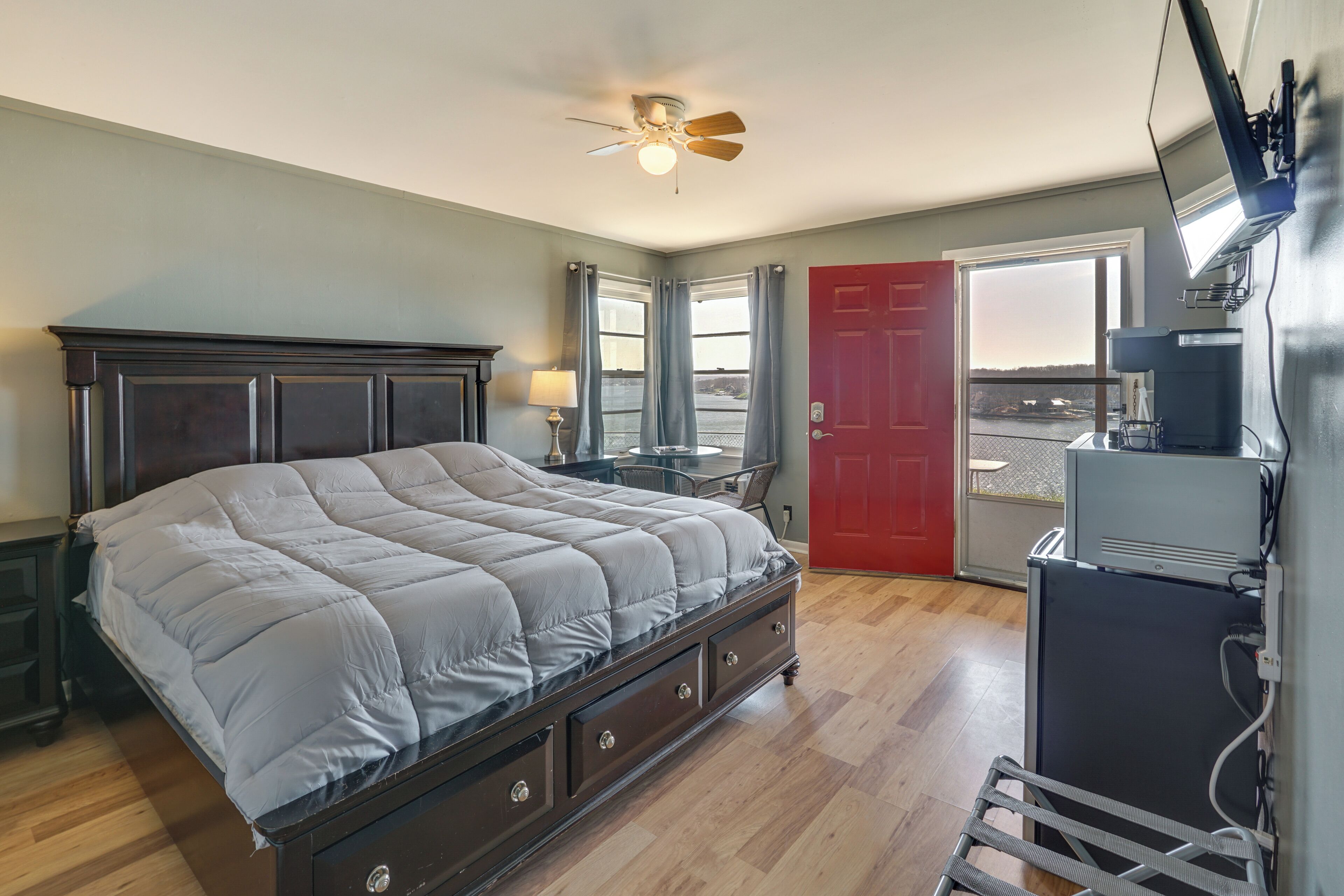 A bedroom with a bed, a ceiling fan, a desk, a chair, a red door, and a view of the outside.