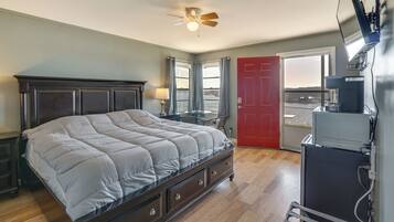 A bedroom with a bed, a ceiling fan, a desk, a chair, a red door, and a view of the outside.