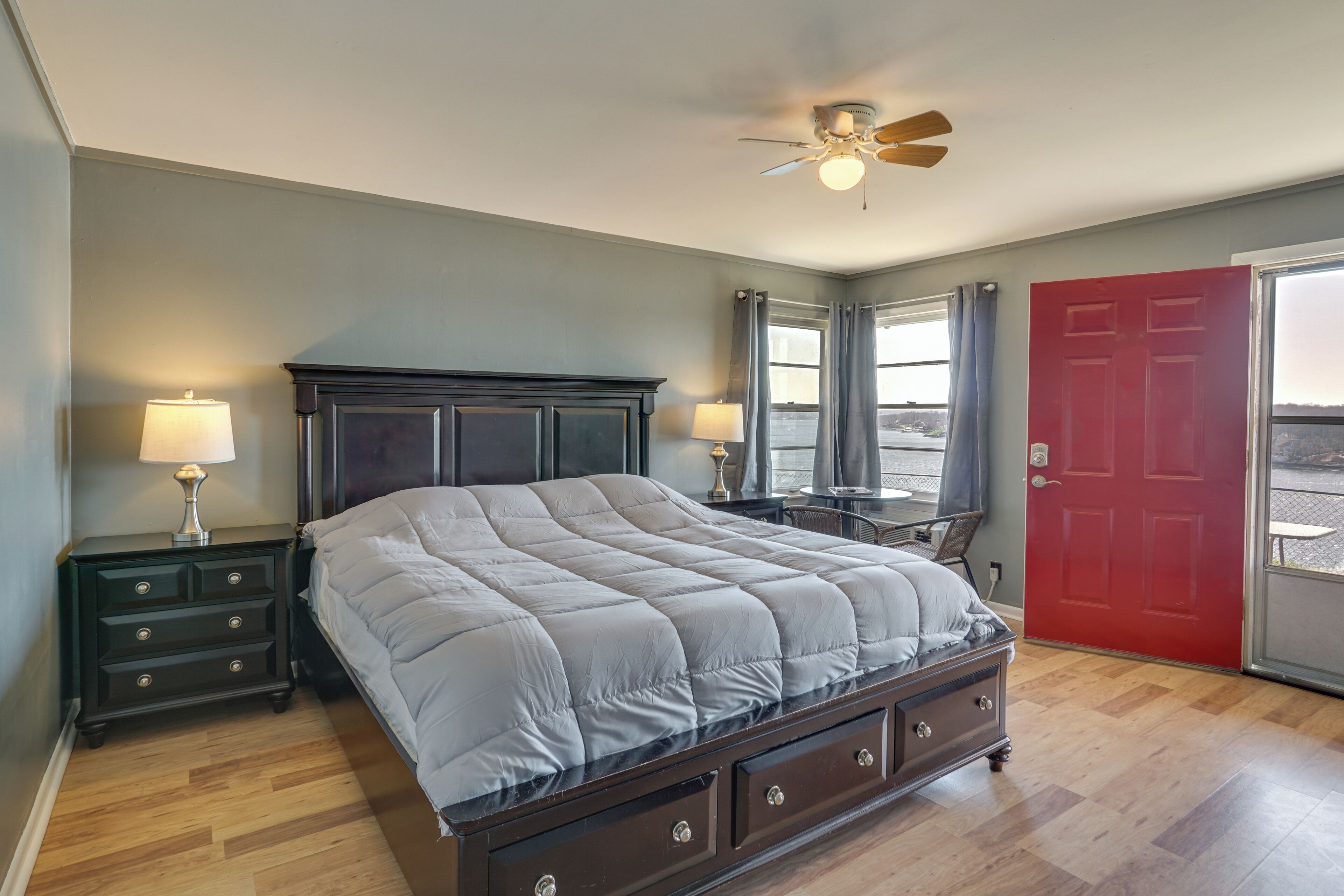 A bedroom with a bed, nightstands, a desk, a red door, and a ceiling fan.