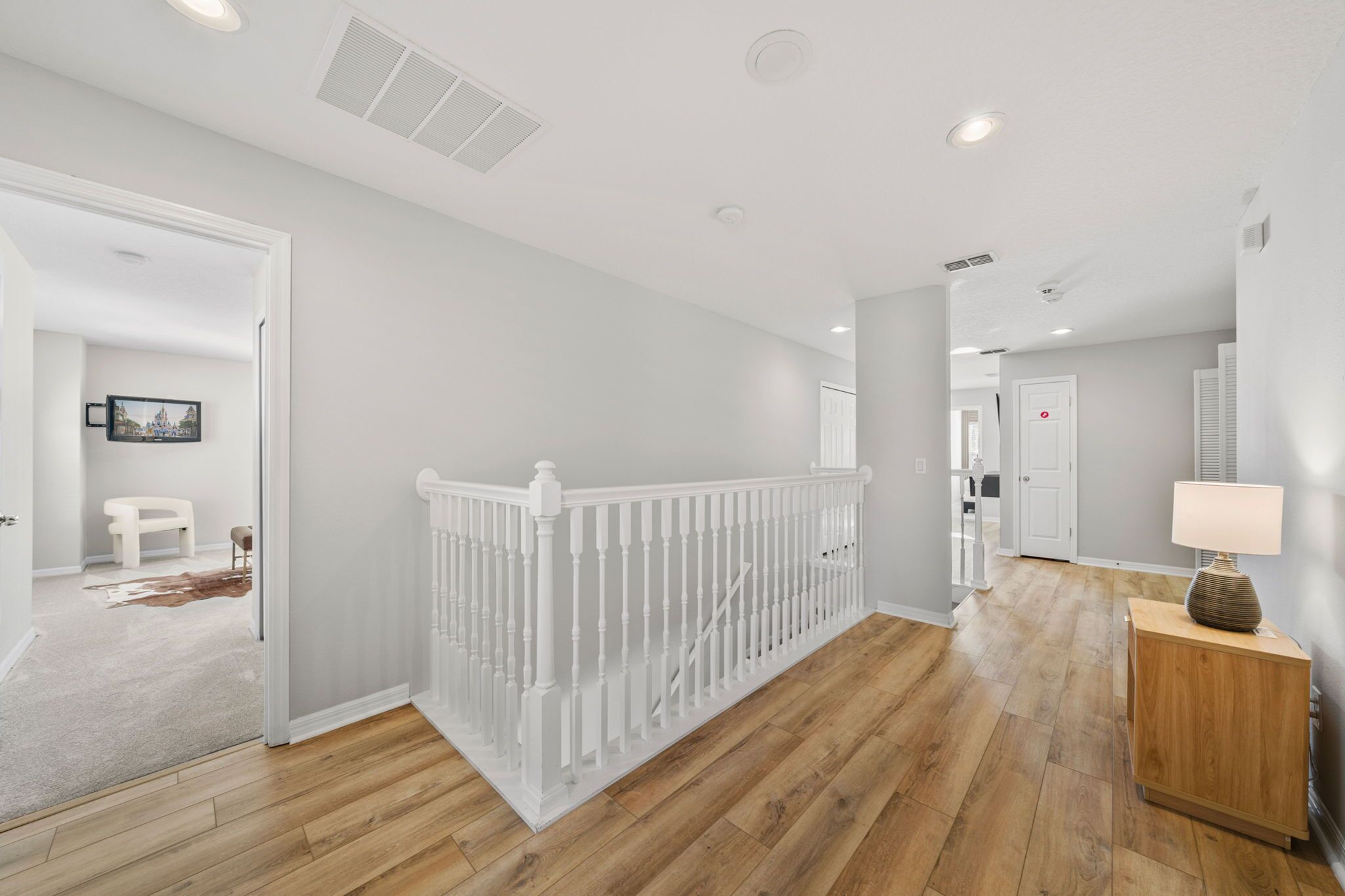 A hallway with a white railing, wooden flooring, and a lamp on a wooden side table.