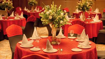 Une salle de banquet avec des tables rondes dressées pour le repas, chacune agrémentée d’un centre de table composé de fleurs, de nappes rouges et de serviettes blanches.