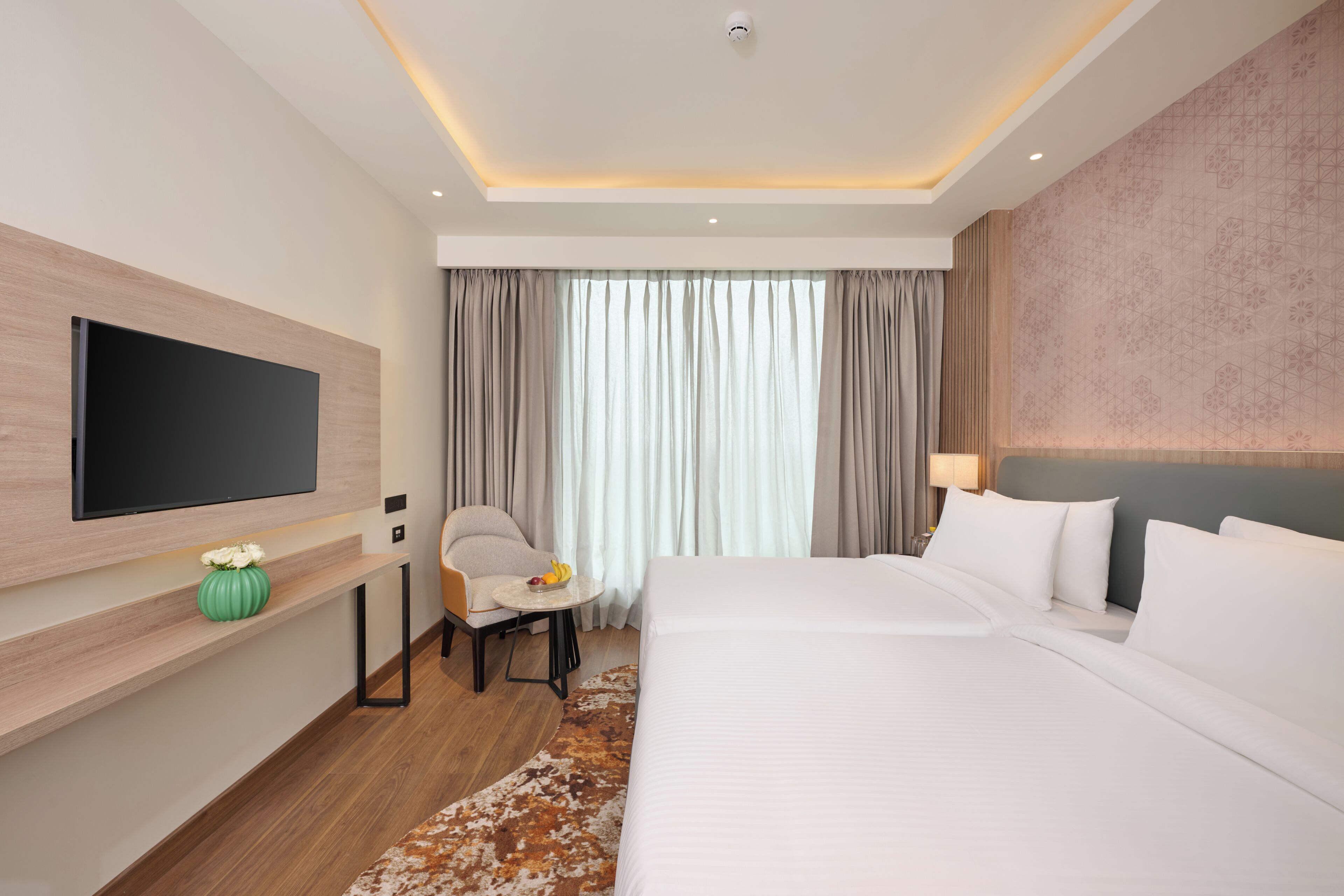 A hotel room with a bed, a TV mounted on the wall, a chair, a small table with fruit, and a window with curtains.