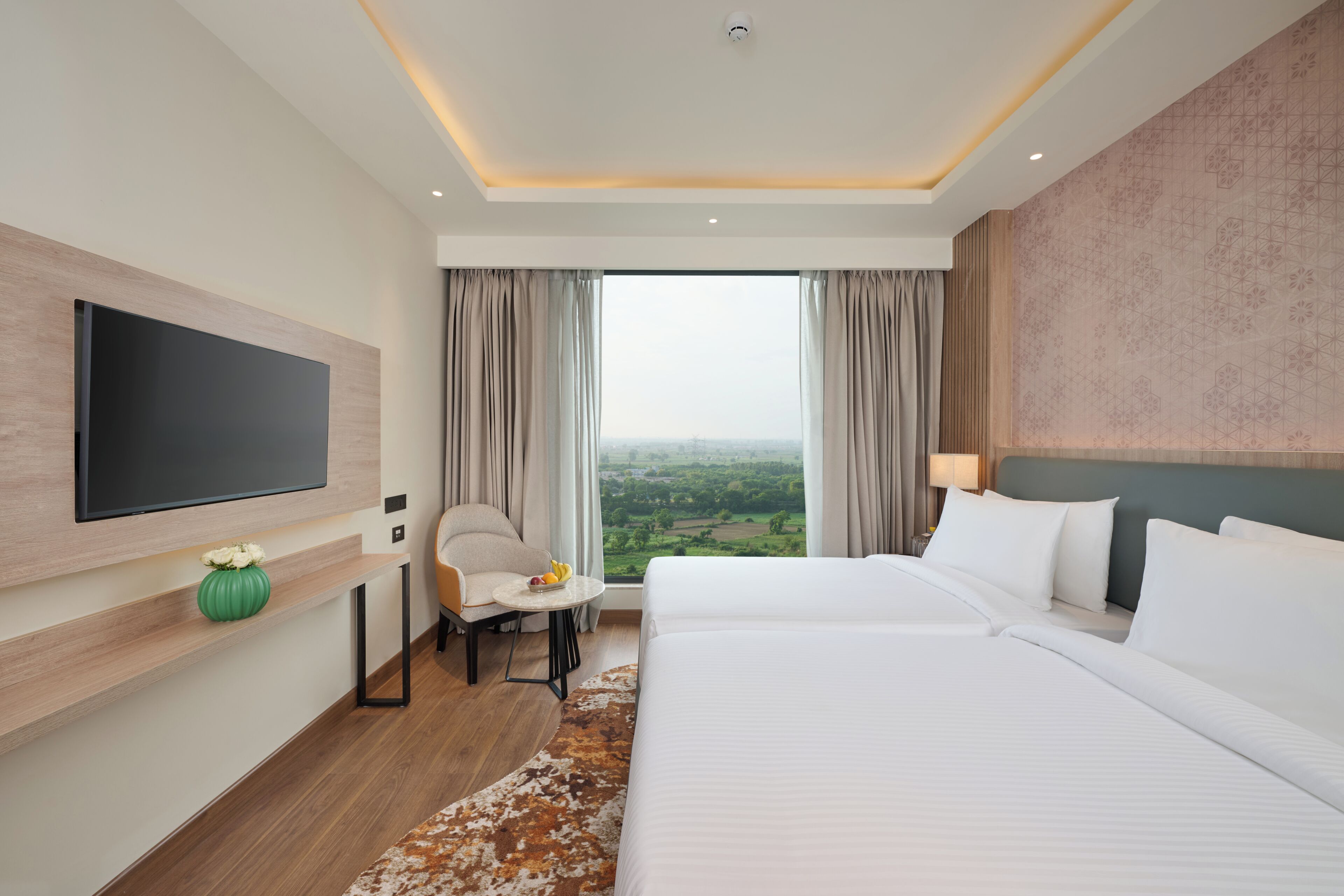 Une chambre d’hôtel avec un grand lit, un téléviseur à écran plat fixé au mur, une petite table avec un saladier de fruits, et une vue sur la campagne par la fenêtre.