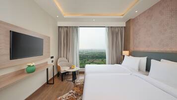 Une chambre d’hôtel avec un grand lit, un téléviseur à écran plat fixé au mur, une petite table avec un saladier de fruits, et une vue sur la campagne par la fenêtre.