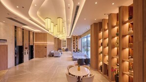 A spacious hotel lobby with a central seating area, a reception desk, and a large window providing a view of the outdoors.