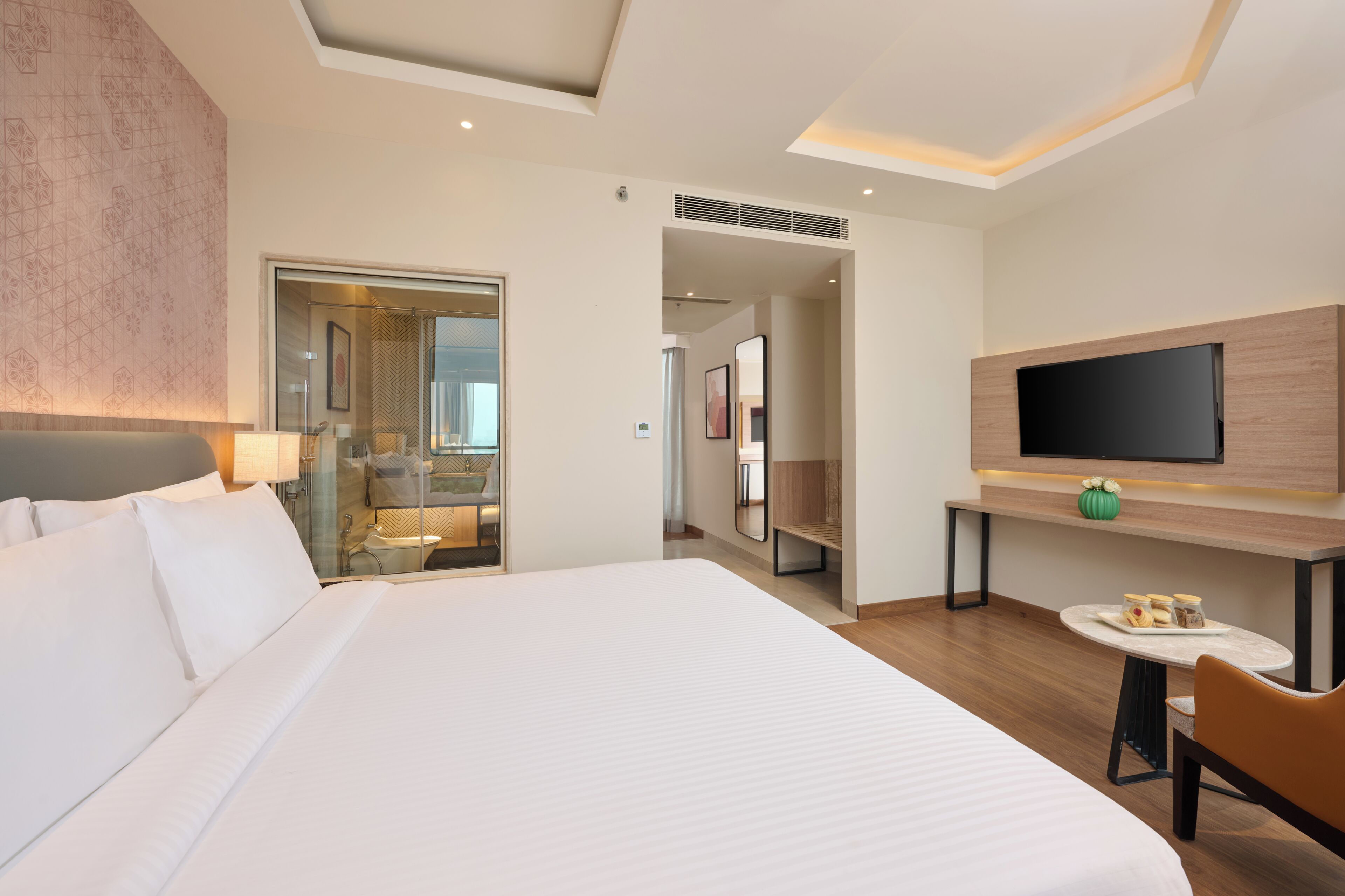 A hotel room with a large bed, a TV mounted on the wall, a small table with a vase, and a bathroom visible through a glass door.