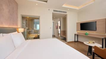 A hotel room with a large bed, a TV mounted on the wall, a small table with a vase, and a bathroom visible through a glass door.