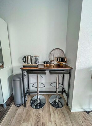 Un coin cuisine avec une table en bois, deux tabourets de bar, une poubelle et un réfrigérateur.