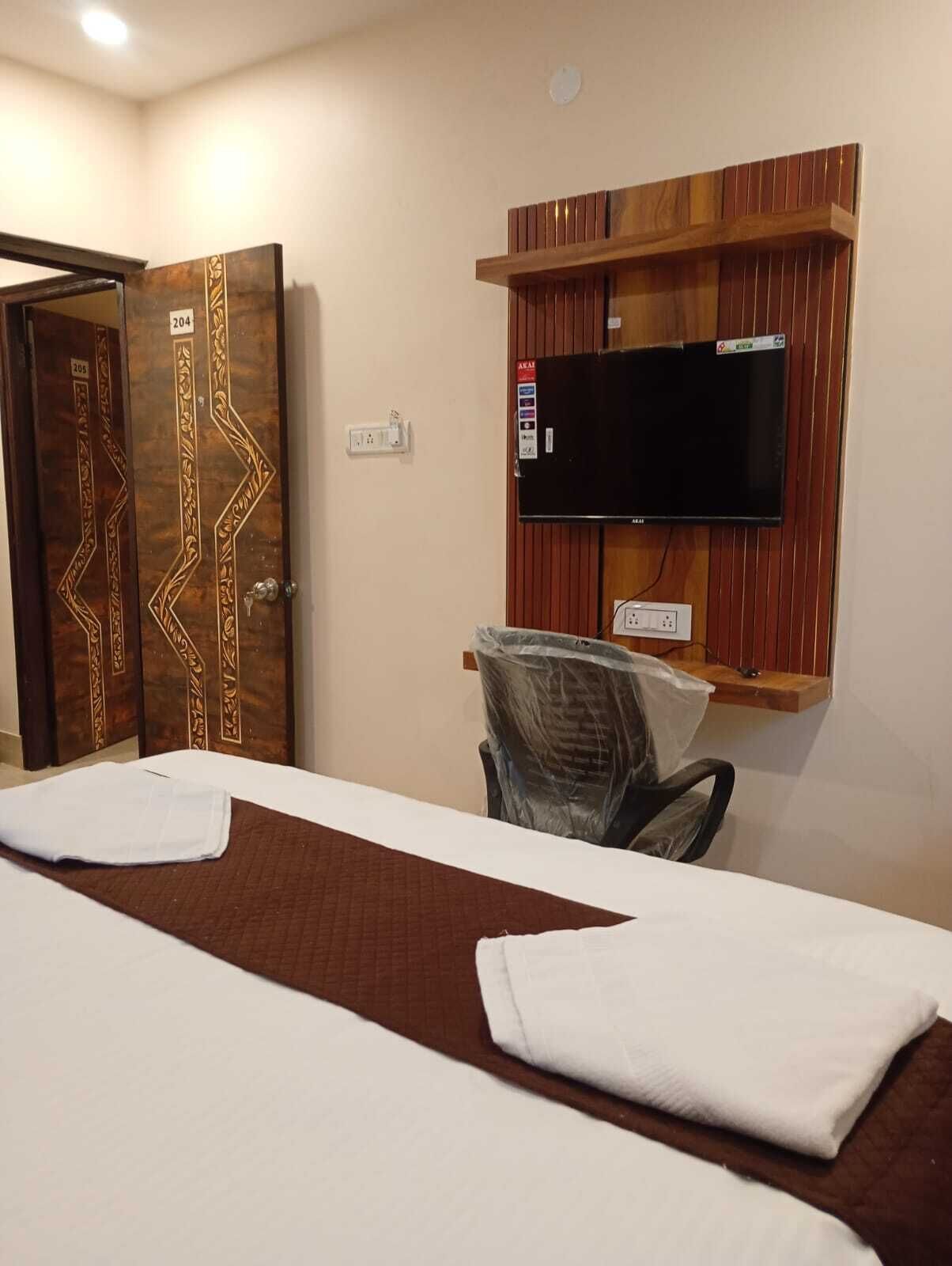 A hotel room with a bed, a television mounted on a wooden panel, a chair with a plastic cover, and a door with decorative patterns.