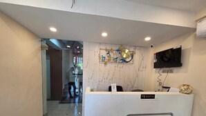 A reception area with a marble wall, a clock, a television, and a sign indicating check-in and check-out times. - The Executive Suites (Barasat)