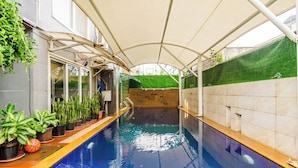 Piscina interior com parede azulejada, plantas em vasos e parede revestida com grama sintética verde.