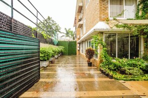 Un étroit sentier humide, bordé de plantes en pot et d’un bâtiment en briques doté d’une clôture verte et d’une porte en verre.