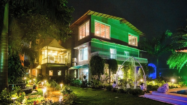 Um edifício de dois andares com varanda, rodeado por vegetação exuberante e um jardim bem iluminado.