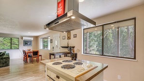 A modern kitchen with a central island, a dining area with a table and chairs, and large windows with a forest view. - Creative Escape! Eagle's Nest Hideaway (Cottage Grove)