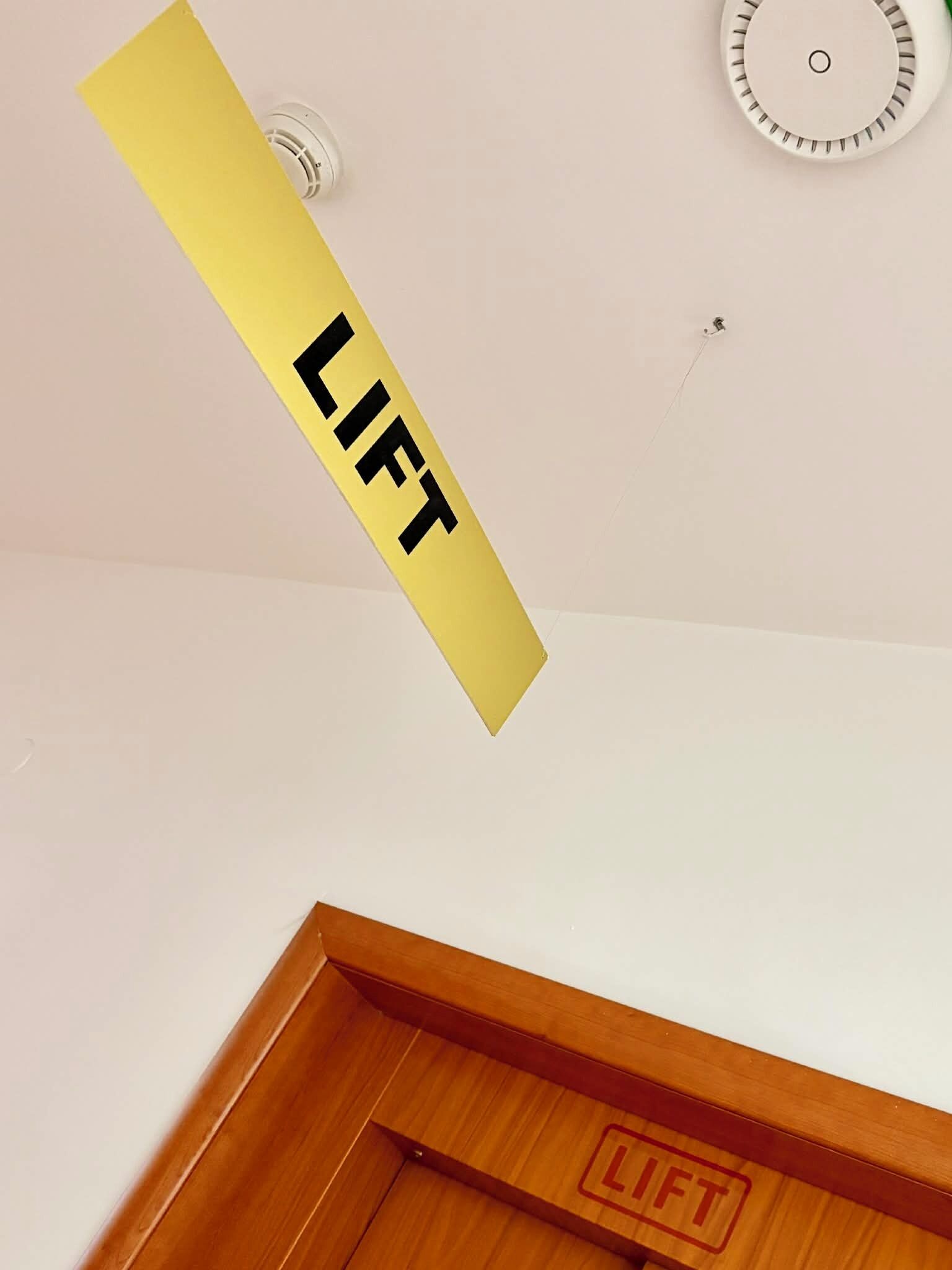 A yellow sign with the word 'LIFT' on a white wall, a ceiling light fixture, and a wooden door frame with a red 'LIFT' symbol.