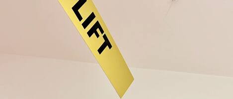 A yellow sign with the word 'LIFT' on a white wall, a ceiling light fixture, and a wooden door frame with a red 'LIFT' symbol.